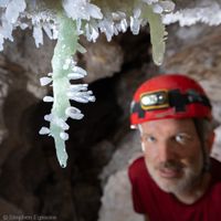 caver in the background looking at a pair of green barite stalactites ornamented with clear calcite crystals